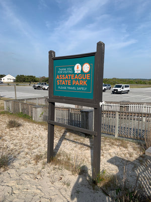 a teal sign with the 5 Tides and Assateague Outfitters logos and orange and white lettering saying "Thank you for visiting Assateague State Park - Please travel safely" framed in dark wood