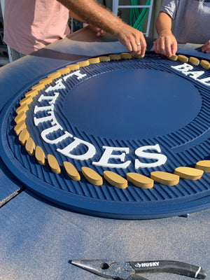 a closeup of a multi-layered sign for Bad Latitudes with two shades of blue and white text, with Wyatt and Justin adding a raised gold rope pattern