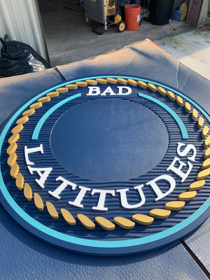 a photo of a multi-layered sign for Bad Latitudes with two shades of blue, a raised gold rope pattern, and white text, missing the logo in the center
