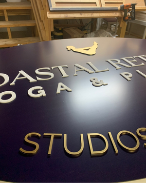 an angled closeup of a large navy blue oval sign with a gold logo and gold and white text for Coastal Retreat Yoga