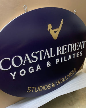 an angled photo of a large navy blue oval sign with a gold logo and gold and white text for Coastal Retreat Yoga