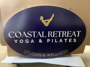 a photo of a large navy blue oval sign with a gold logo and gold and white text for Coastal Retreat Yoga