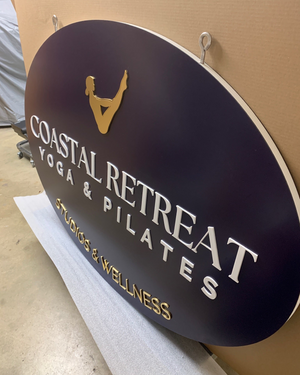 an angled photo of a large navy blue oval sign with a gold logo and gold and white text for Coastal Retreat Yoga