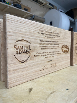 an angled photo of a wooden plank laser engraved with the Samuel Adams and Dogfish Head logos and text