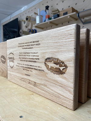 an angled photo of a wooden plank laser engraved with the Samuel Adams and Dogfish Head logos and text