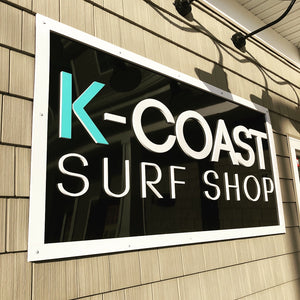a photo of a K-Coast Surf Shop sign with a white border and lettering on a black background