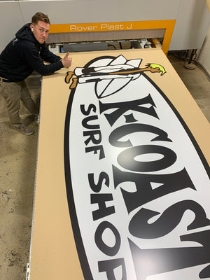 a photo of a large K-Coast Surf Shop logo on wood in the shop, and Justin giving a thumbs up