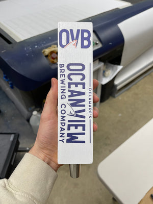 a white rectangular wooden tap handle with Ocean View Brewing Company's name and logo printed in navy blue