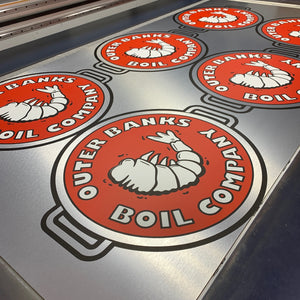 a photo of six of the Outer Banks Boil Company logos printed on a sheet of uncut aluminum